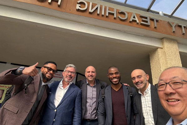 A group of men smiling for a selfie outside a building with a sign that includes the word Teaching.