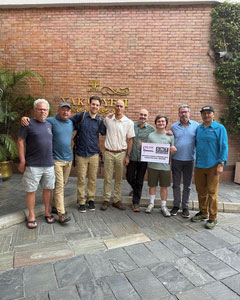 Group of eight men posing together outside a brick building entrance, with one holding a small sign.