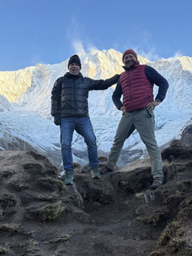Two men in winter gear standing on a rocky trail with snowy peaks behind them.