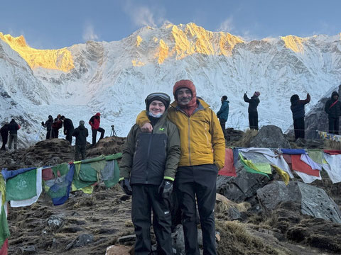 Two trekkers posing together in front of a massive, sunlit snow-covered mountain range.