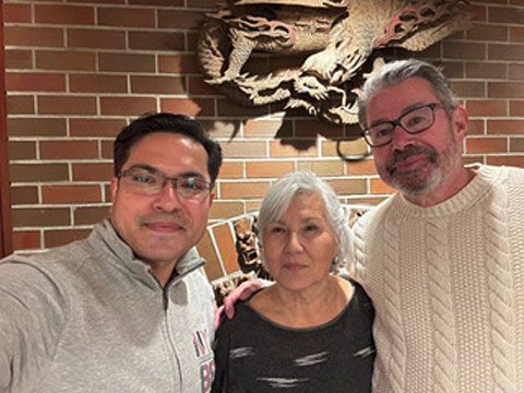 A woman flanked by two men smiling together in front of a brick fireplace.