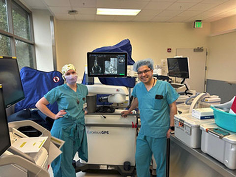 Two medical professionals in scrubs posing next to an ExcelsiusGPS robotic navigation system.