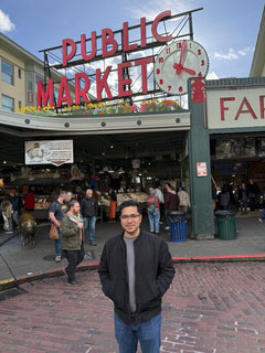 A man posing outside on a brick walkway beneath a large Public Market clock sign.