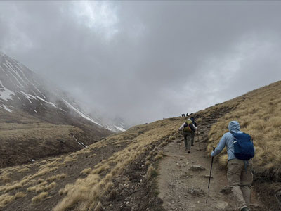 Trekkers hiking up a dirt trail on a grassy mountain slope under a cloudy sky.
