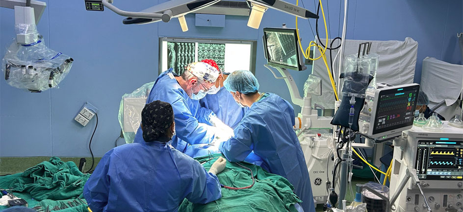 A team of surgeons in blue gowns and surgical caps performing a procedure in an operating room filled with medical monitors and equipment.