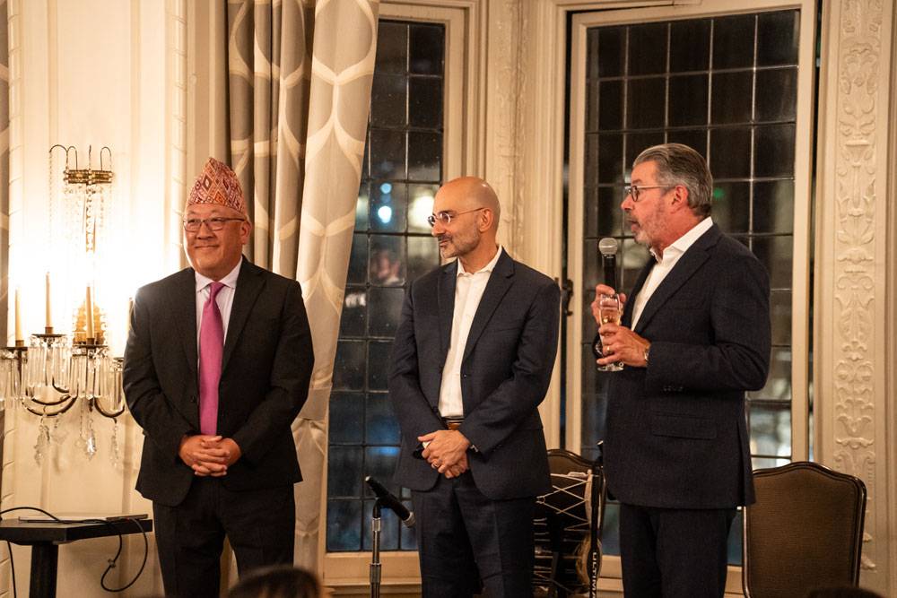 Three men in formal attire speaking at a gala; one wears a traditional Nepali Dhaka topi.