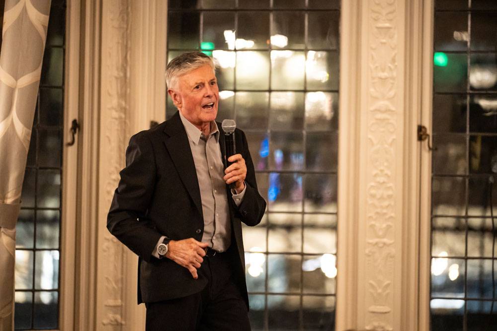 An older man in a black blazer speaks enthusiastically into a microphone during an event.