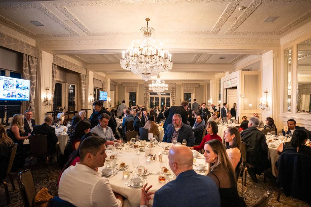 Wide view of a crowded formal banquet hall with guests seated at round tables under large chandeliers.
