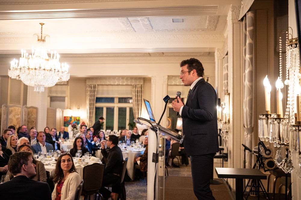 A man in a suit gives a presentation from a podium to a seated audience in a formal ballroom.
